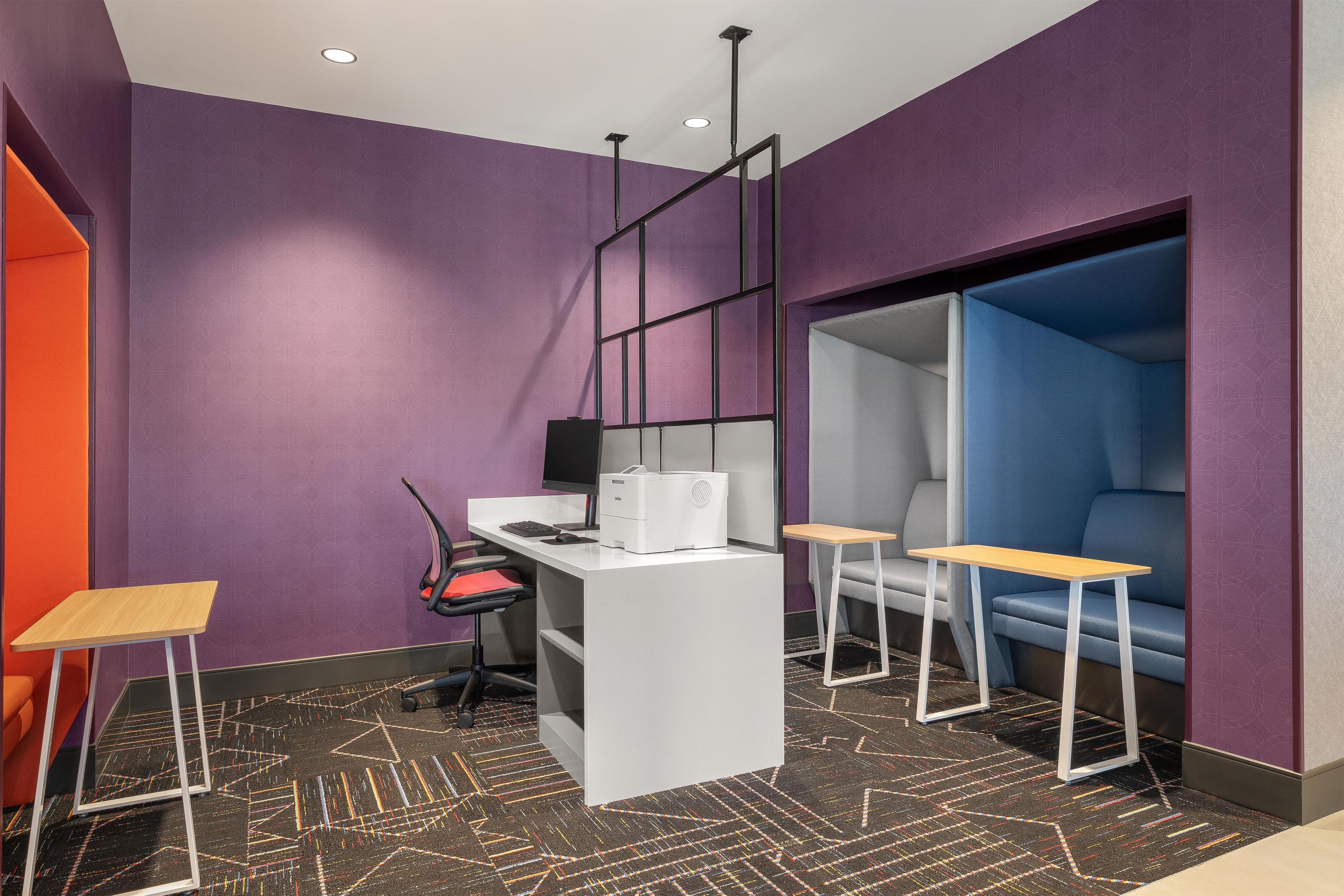 A modern office space with a white desk, a computer, and a printer. There are two chairs and a small table. The walls are purple, and the floor has a geometric patterned carpet.