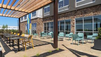 An outdoor patio with tables and chairs, a pergola, and a stone building with large windows.