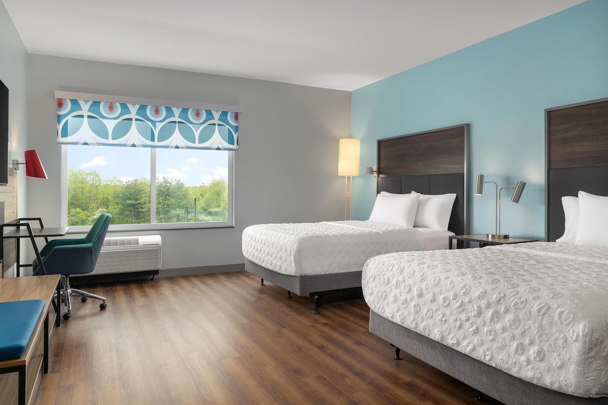 A hotel room with two beds, a desk, a chair, a window with a view, and a wall-mounted lamp.