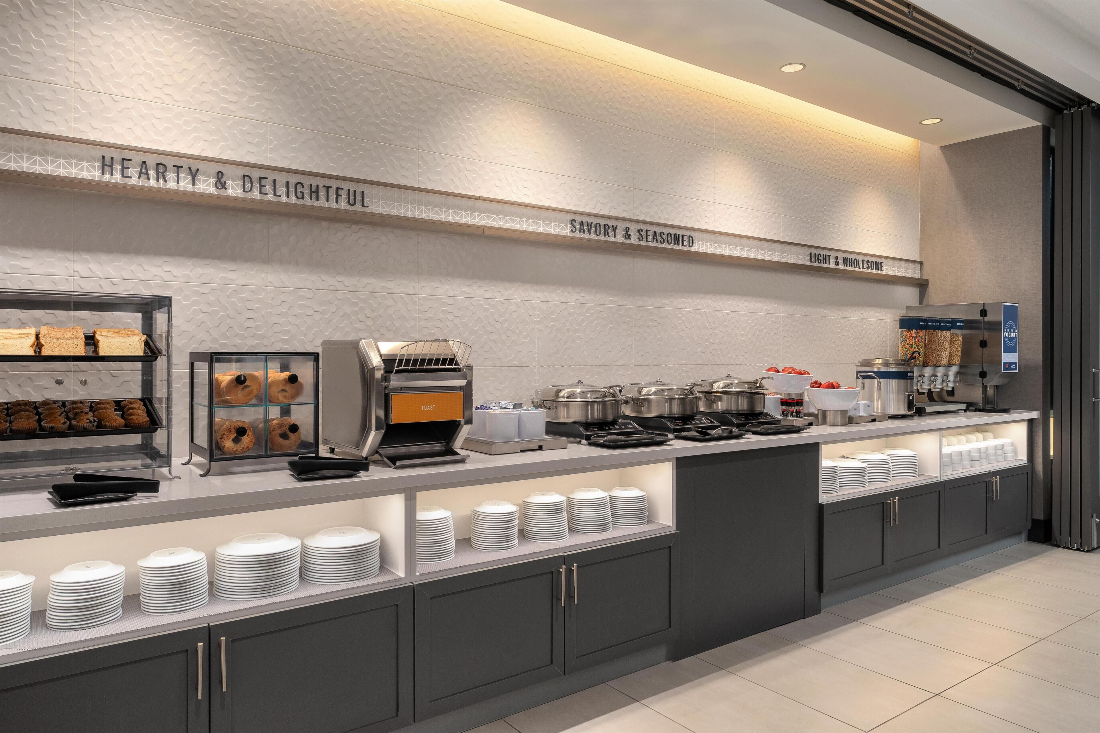 A modern breakfast bar with labeled sections for different food categories, a coffee machine, and neatly arranged plates and bowls.