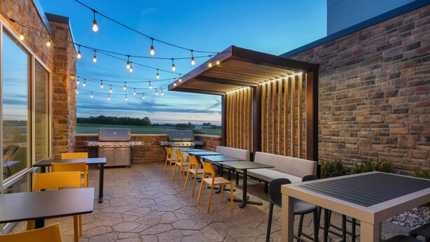 An outdoor patio with stone flooring, wooden tables and chairs, string lights, and a grill area. - Tru By Hilton Burlington, Nj (Burlington)