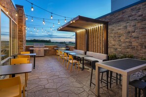 An outdoor patio with stone flooring, wooden tables and chairs, string lights, and a grill area.
