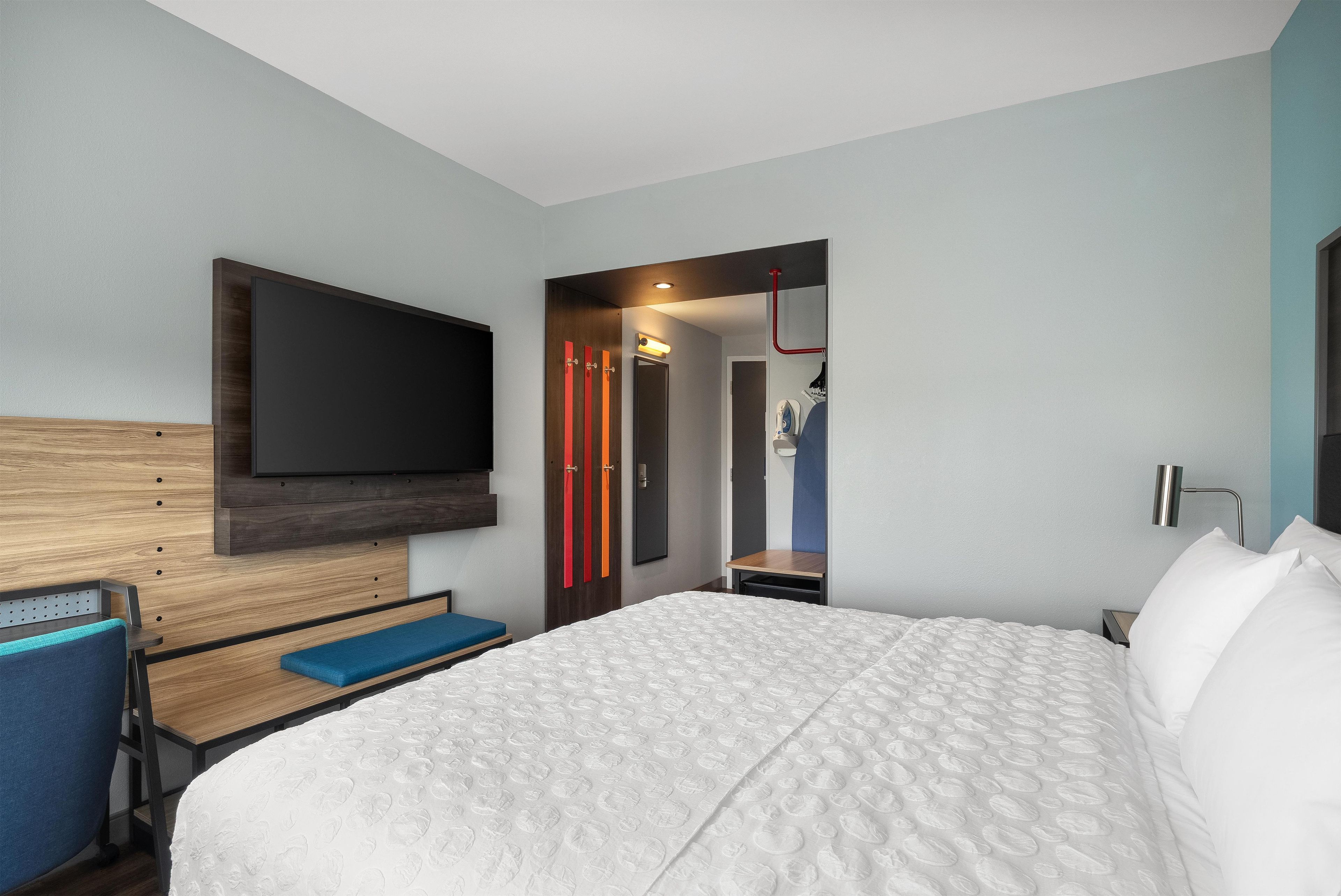 A modern hotel room with a large bed, a desk with a chair, a flat-screen TV mounted on the wall, and a walk-in closet with a mirror.