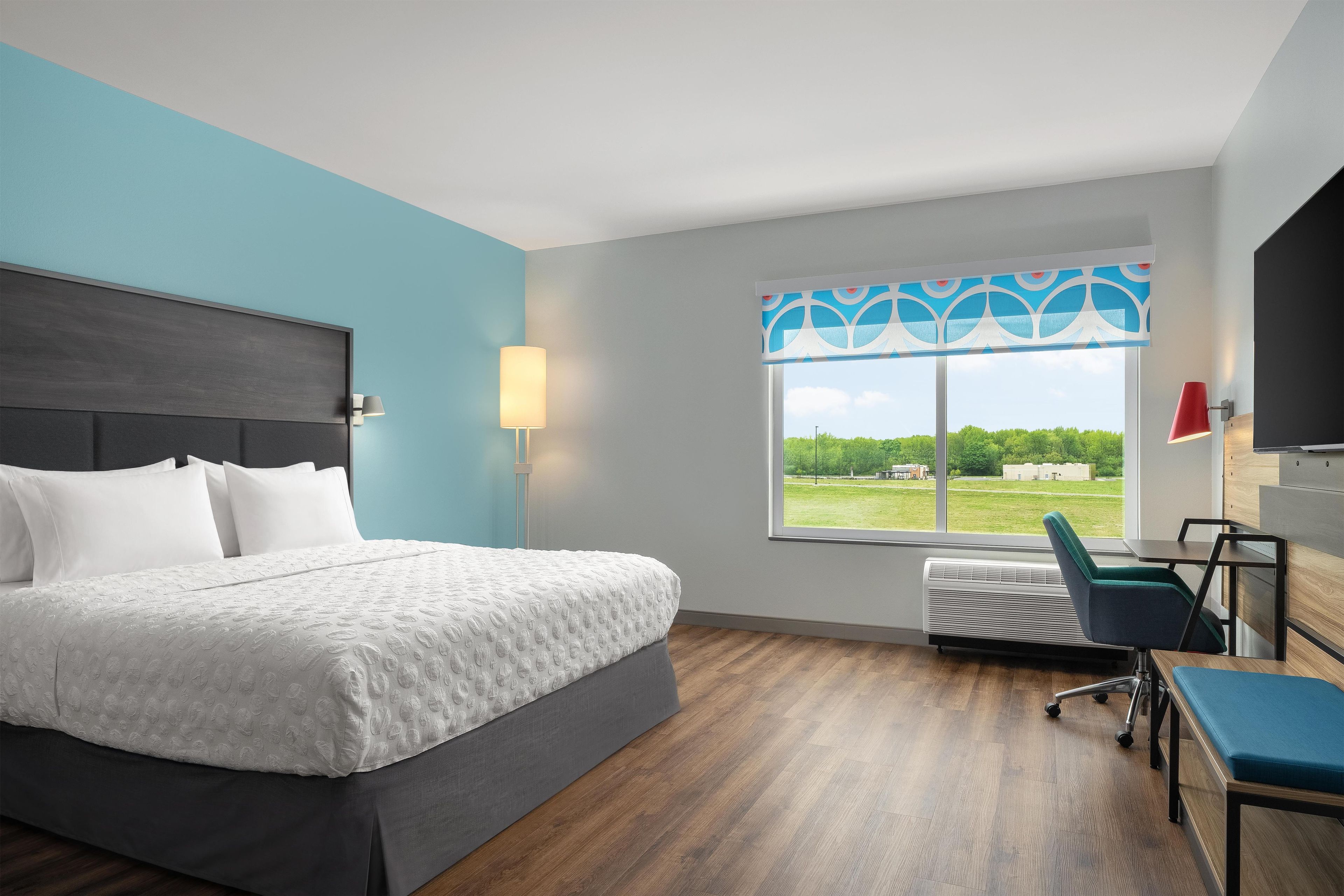A modern hotel room with a large bed, a desk, a chair, a window with a view, and a wall-mounted TV.