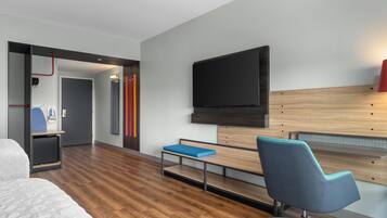 A modern hotel room with a wooden floor, a bed, a desk with a chair, a TV mounted on the wall, and a small seating area with a bench.