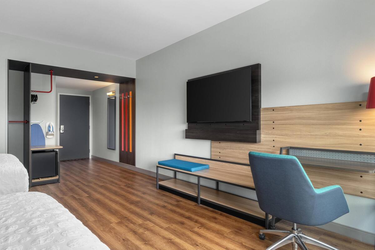 Una habitación de hotel moderna con suelo de madera, una cama, un escritorio con una silla, un televisor montado en la pared y una pequeña zona de estar con un banco.