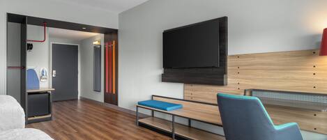 A modern hotel room with a wooden floor, a bed, a desk with a chair, a TV mounted on the wall, and a small seating area with a bench.