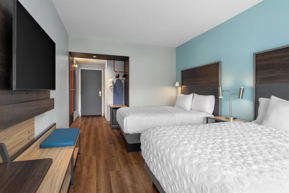 Habitación de hotel con dos camas, televisión, banco y suelo de madera.