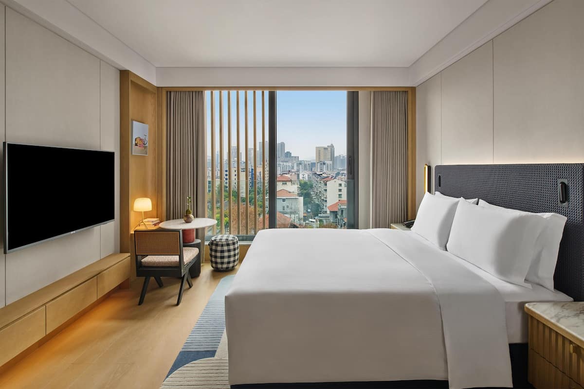A hotel room with a large bed, a TV, a small table with a lamp, a chair, and a view of the city.
