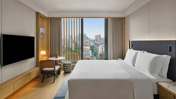 Habitación de hotel con una cama grande, televisión, una mesita con lámpara, una silla y vistas a la ciudad.
