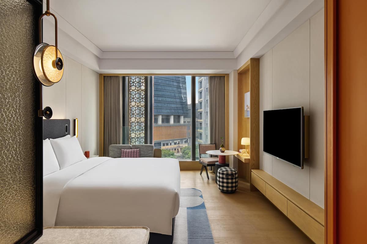 A hotel room with a large bed, a flat-screen TV, a small table, and a view of the cityscape through large windows.