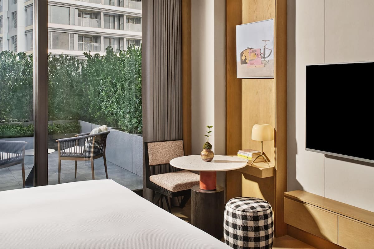 A modern hotel room with a large window, a round table with a plant, a sofa, a television, and a small cabinet.