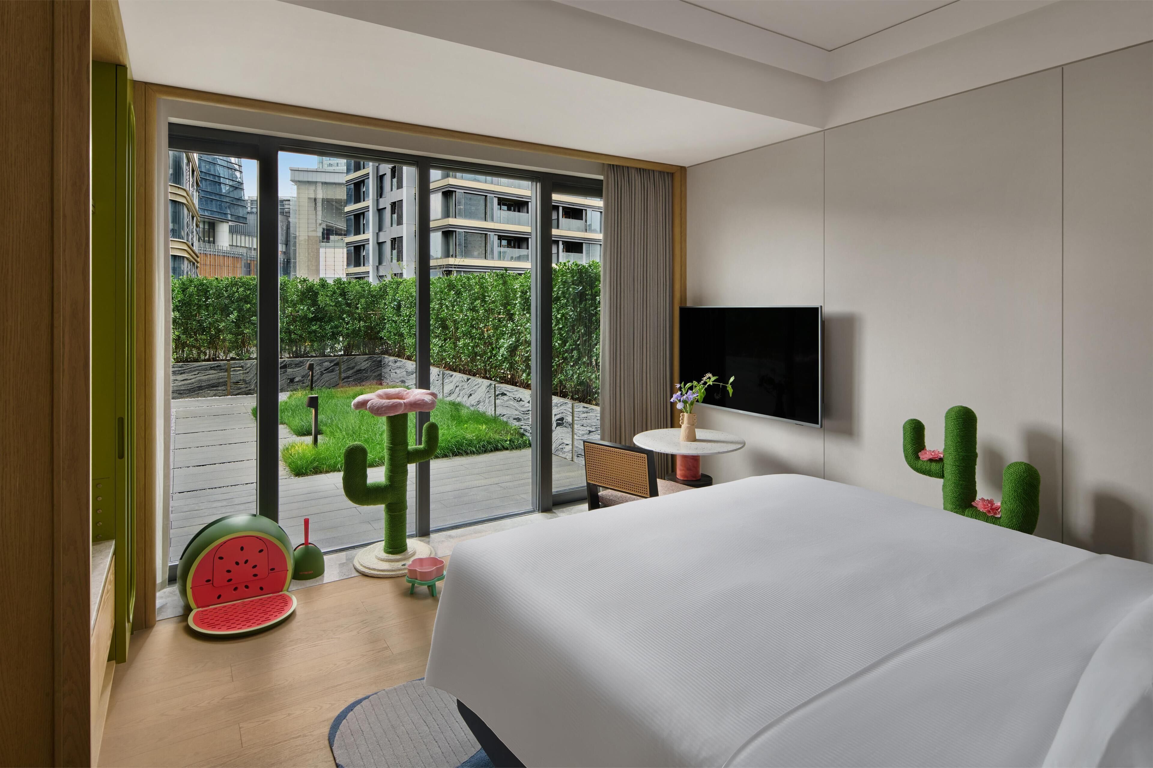 Una habitación de hotel moderna con una cama grande, un televisor, una mesita con una planta y vistas de la ciudad a través de ventanas amplias.