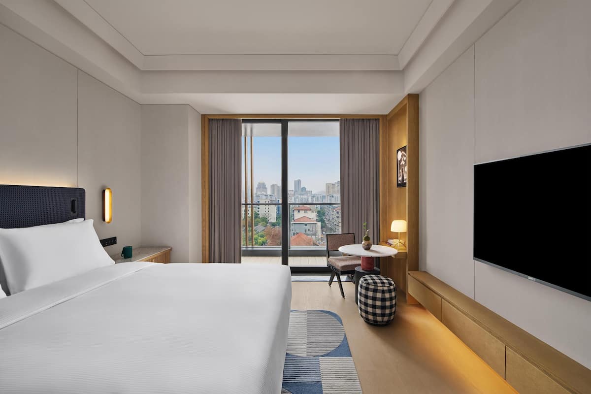 A hotel room with a bed, a desk, a chair, a TV, and a large window with a city view.