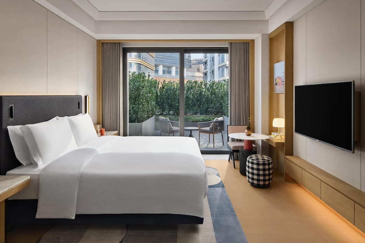 A hotel room with a large bed, a TV, a balcony with a view of buildings, and a small table with chairs.