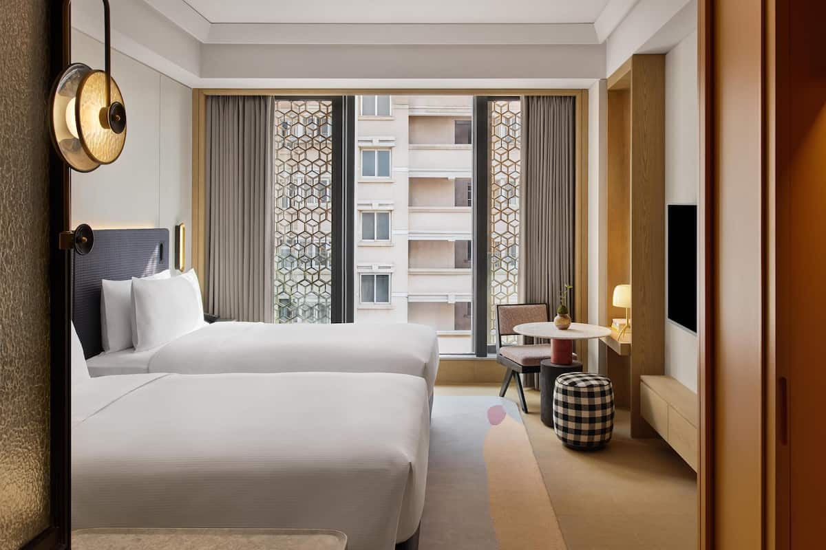 A hotel room with two beds, a TV, a small table with a chair, and a window with a view of buildings.