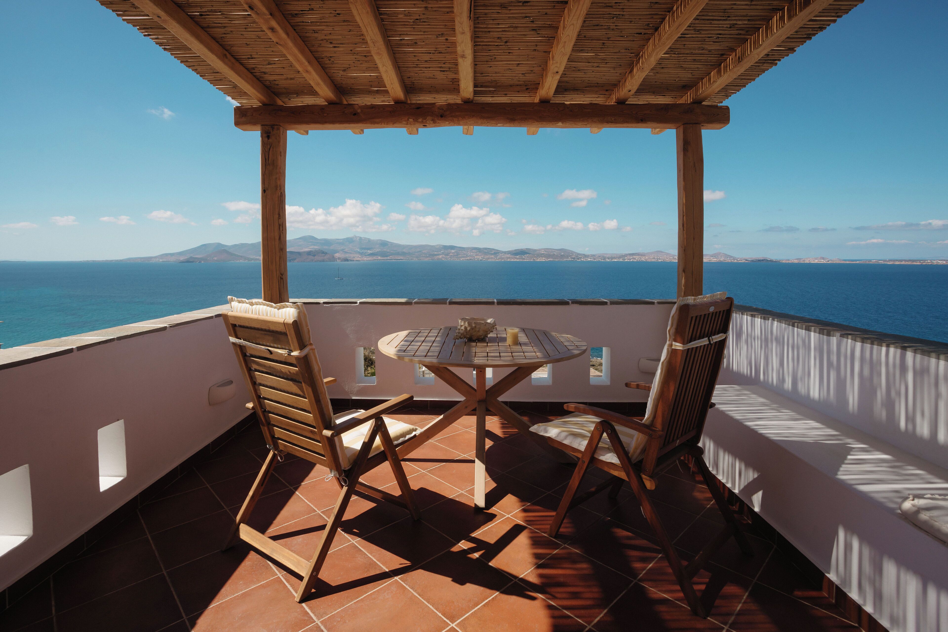 A terrace with a wooden table and chairs, overlooking a body of water and distant mountains.