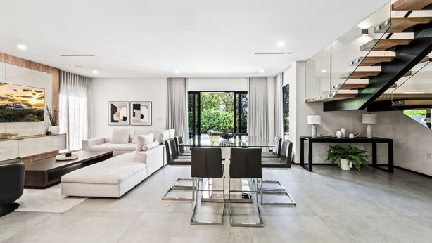 A modern living room with a sofa, dining table, and chairs, a staircase, and a view of greenery outside. - Ocean Crown Villa - A Modern Beach Getaway (Fort Lauderdale)