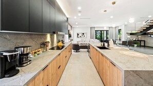 A modern kitchen with dark cabinets, a central island, and a dining area with a staircase. - Ocean Crown Villa - A Modern Beach Getaway (Fort Lauderdale)
