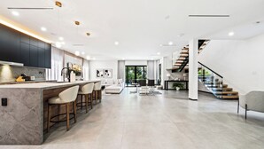 A spacious modern kitchen with a central island, bar stools, and a dining area with a sofa and chairs. - Ocean Crown Villa - A Modern Beach Getaway (Fort Lauderdale)