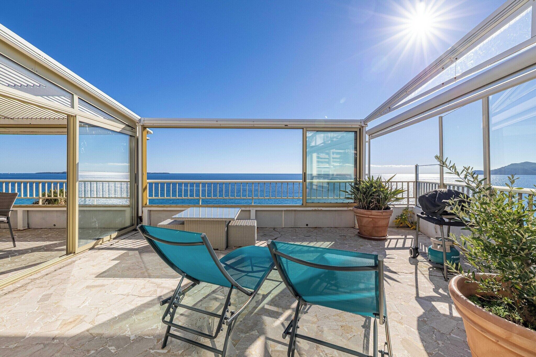 Un espace extérieur ensoleillé avec vue sur la mer, comprenant une table, des chaises et des plantes en pot.