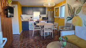 A compact kitchen with dark cabinets, a dining area with wicker chairs, and a living space with a sofa and a plant. - The yellow house (Vidauban)