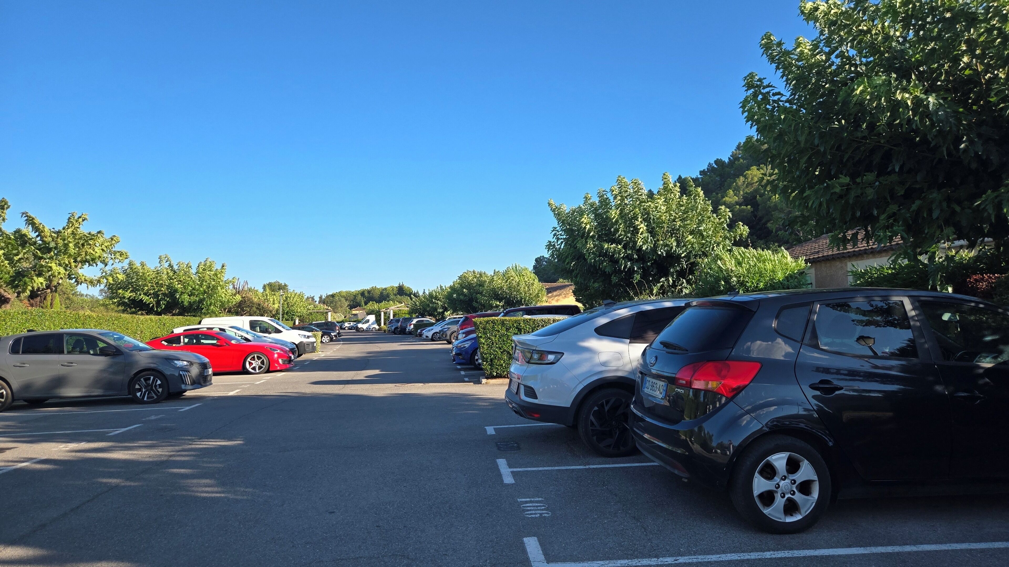 A parking lot with multiple cars, including a red sedan and a white SUV, with trees and a clear blue sky in the background.