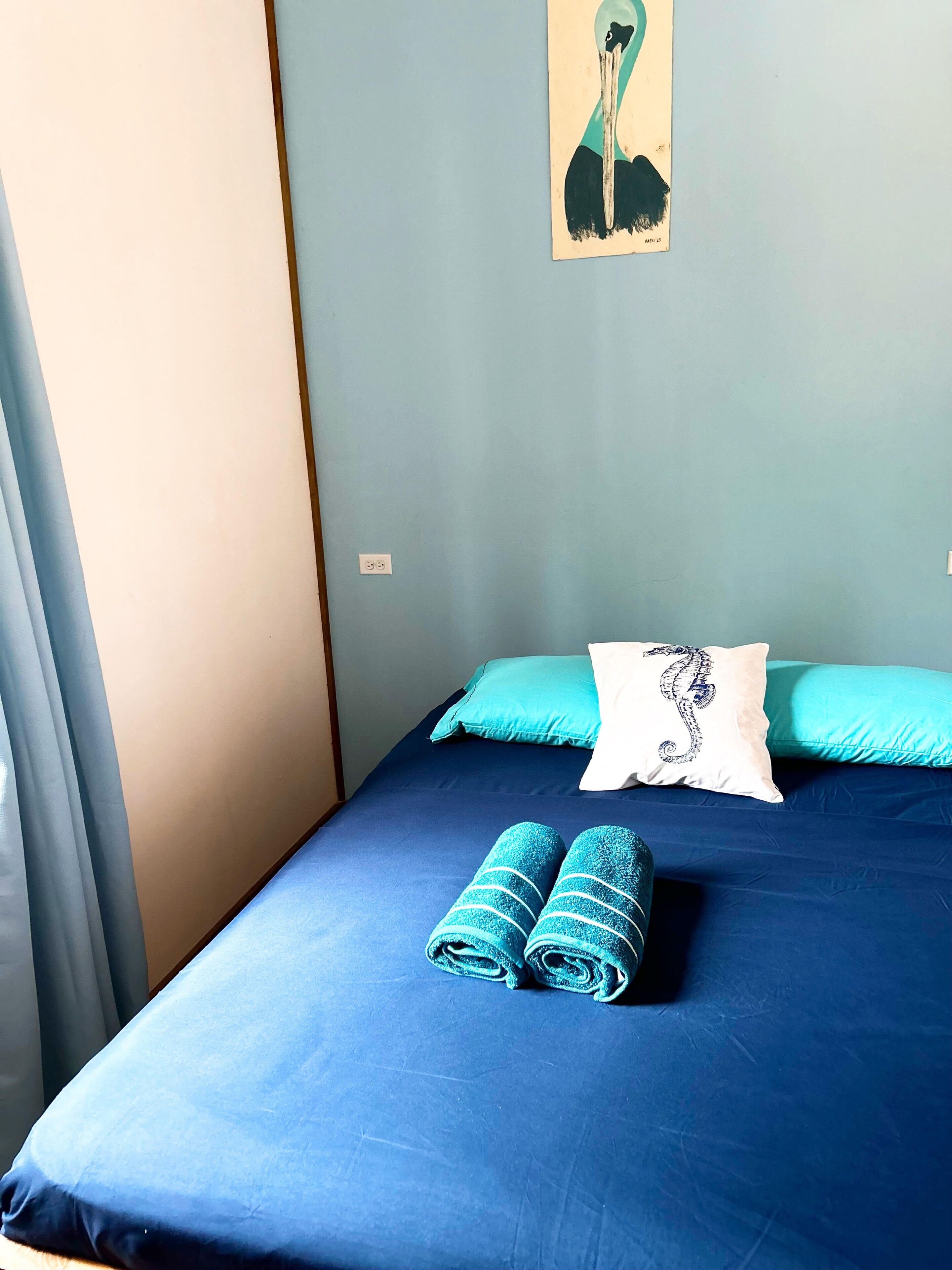 Una cama con colcha azul, dos toallas enrolladas y un almohadón con diseño de caballito de mar.