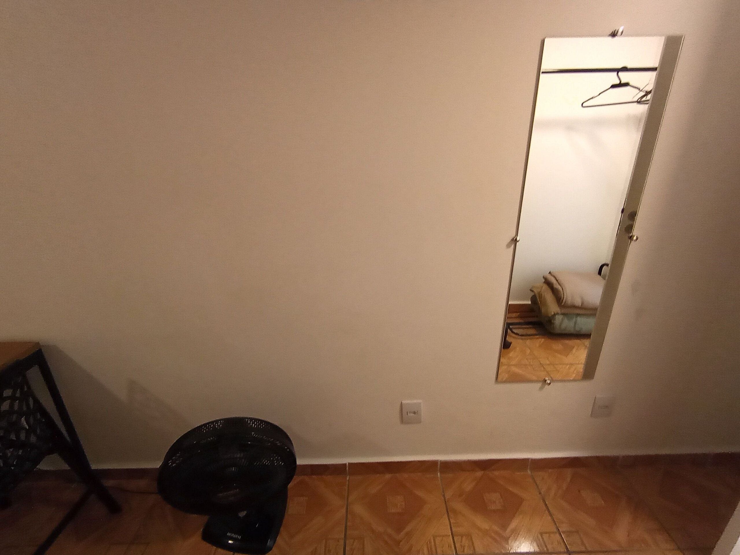 Een kamer met een houten vloer, een zwarte prullenbak, een stoel en een spiegel die een bed en een hangende jas weerspiegelt.
