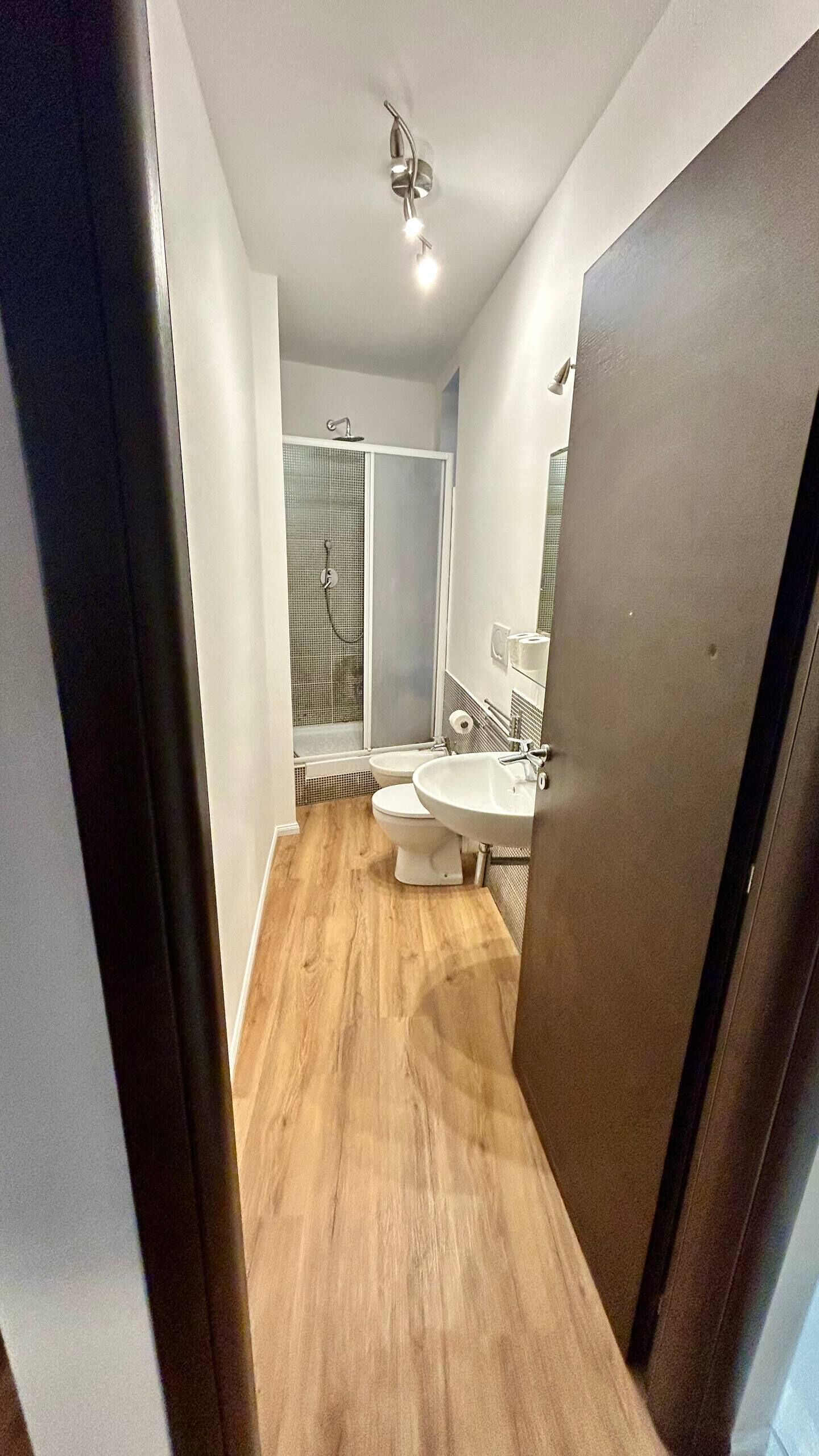 Un couloir étroit mène à une salle de bain équipée d’une douche, de deux lavabos et de toilettes.