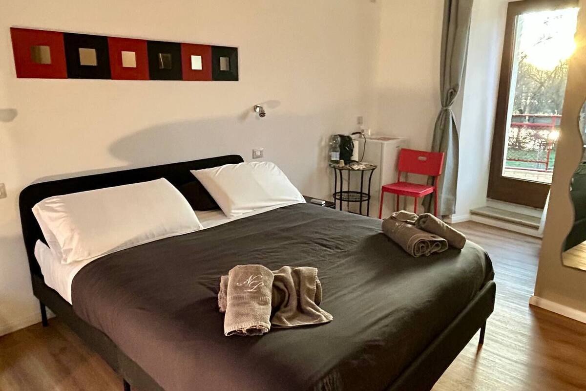 Una cama bien tendida con un cabecero oscuro, una mesita con una silla y una ventana con cortinas.