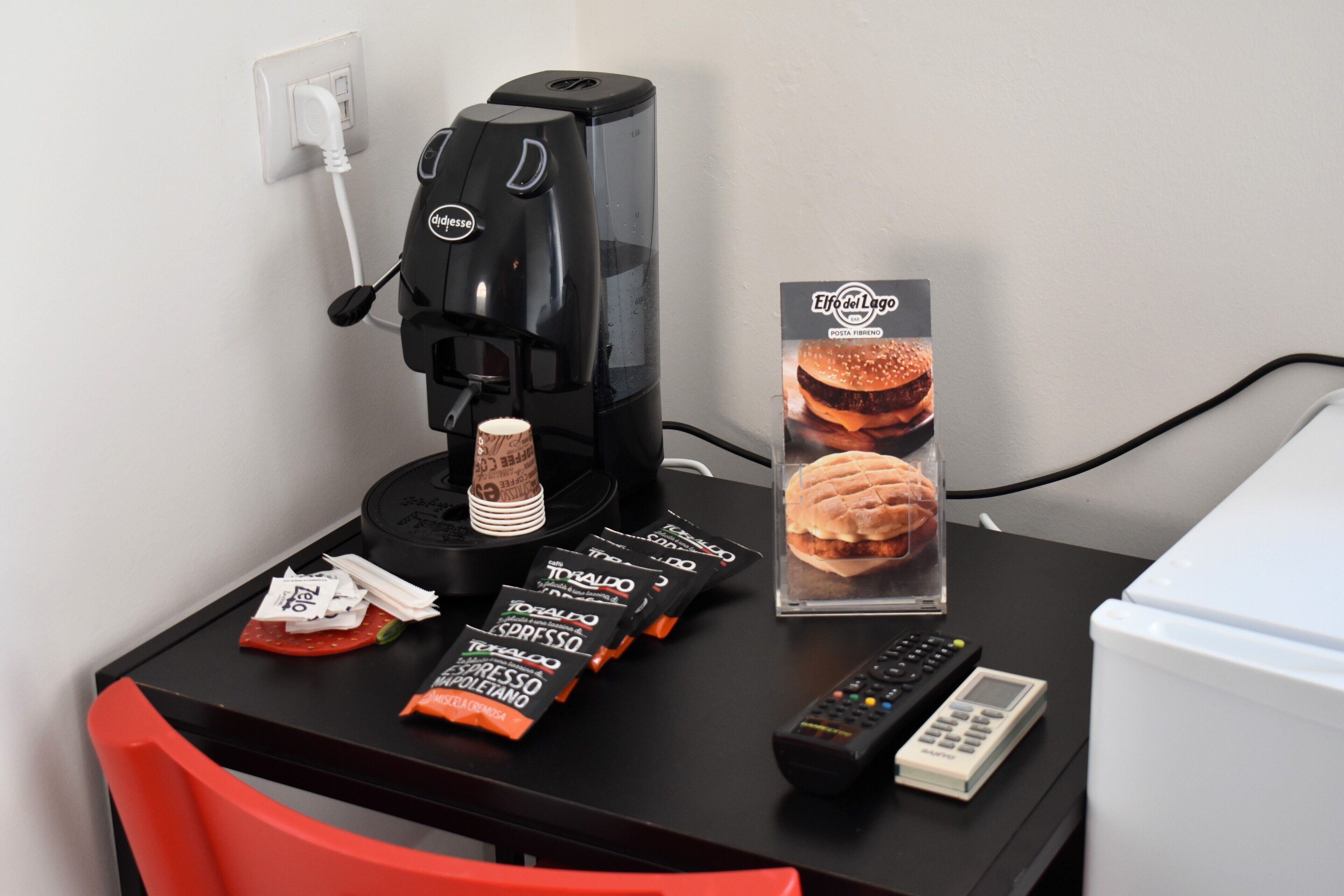 A small table with a coffee maker, a sandwich, a remote control, and some coffee packets.
