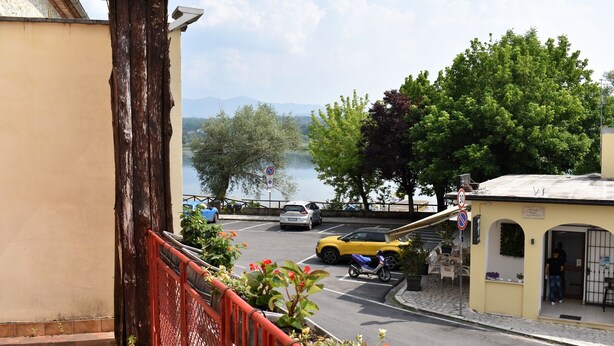 A street view with a yellow car parked by the roadside, a small building with a sign, and a view of a lake with trees in the background. - Ninfa del Lago Bedrooms  (Posta Fibreno)