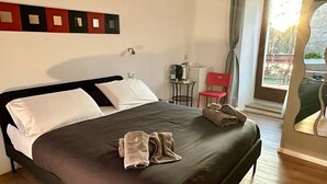A neatly made bed with a dark bedspread and pillows, a small table with a chair, and a window with curtains. - Ninfa del Lago Bedrooms  (Posta Fibreno)