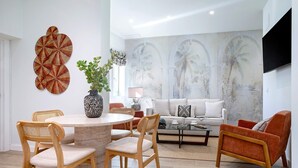 Una sala moderna con una mesa de comedor redonda, dos sillas de madera, un sofá y una mesita de centro. Hay un mural grande con palmeras y un televisor de pantalla plana montado en la pared.