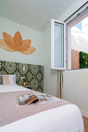 Un dormitorio con un cabecero estampado, una cama con una revista, una ventana con vistas y una lámpara fijada a la pared.