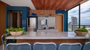 A modern kitchen with a built-in refrigerator, a sink, and a wooden ceiling. There are two potted plants on the counter and a view of the city through large windows. - Fully Equipped Accommodations in a Building With a Pool, 270m From Barra Beach - Caoa (Salvador)
