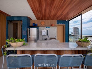 A modern kitchen with a built-in refrigerator, a sink, and a wooden ceiling. There are two potted plants on the counter and a view of the city through large windows. - Fully Equipped Accommodations in a Building With a Pool, 270m From Barra Beach - Caoa (Salvador)