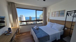 A hotel room with a large window offering a city view, a bed with a blue bedspread, a desk with a chair, and a small table with a chair. - Fully Equipped Accommodations in a Building With a Pool, 270m From Barra Beach - Caoa (Salvador)