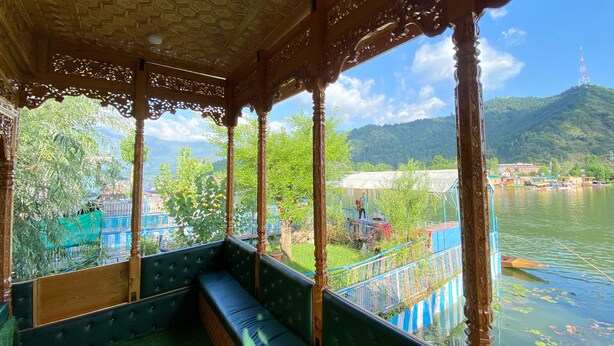 A covered seating area with a bench, wooden pillars, and a view of a river with boats and a distant hill. - The Apollo 11 Heritage Luxury Houseboat (Srinagar)