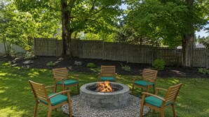 A circular stone fire pit surrounded by six wooden chairs with green cushions, set in a grassy backyard with a wooden fence in the background. - Quaint Bethel Home with Firepit - Walk to Town (Bethel)