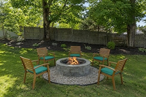 A circular stone fire pit surrounded by six wooden chairs with green cushions, set in a grassy backyard with a wooden fence in the background. - Quaint Bethel Home with Firepit - Walk to Town (Bethel)