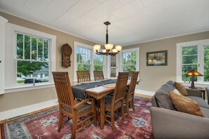A dining area with a wooden table and chairs, a chandelier, and large windows with a view of trees. - Quaint Bethel Home with Firepit - Walk to Town (Bethel)