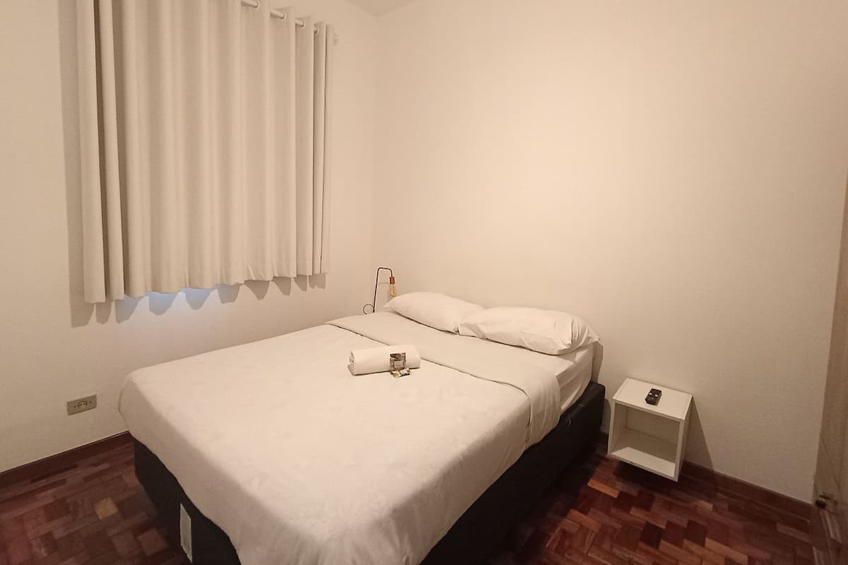 A single bed with a white mattress and pillows, a bedside table with a lamp, and a window with curtains.