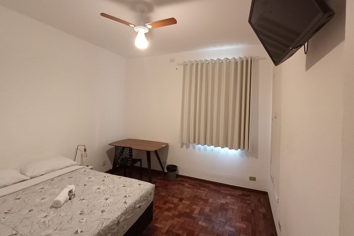 A bedroom with a bed, a ceiling fan, a TV, a window with blinds, a small table, and a chair.