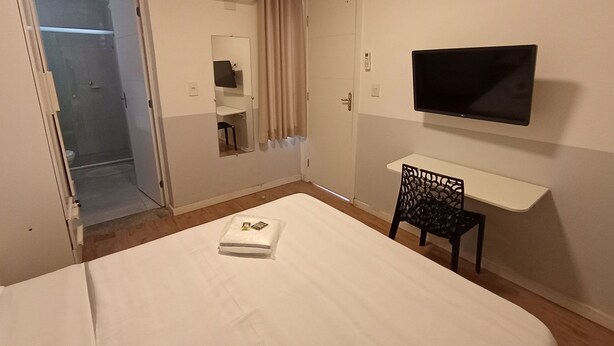 A hotel room with a bed, a TV, a desk, a chair, a mirror, a shower, and an air conditioning unit. - WeStay Budget-Unidade 3 (São Paulo)