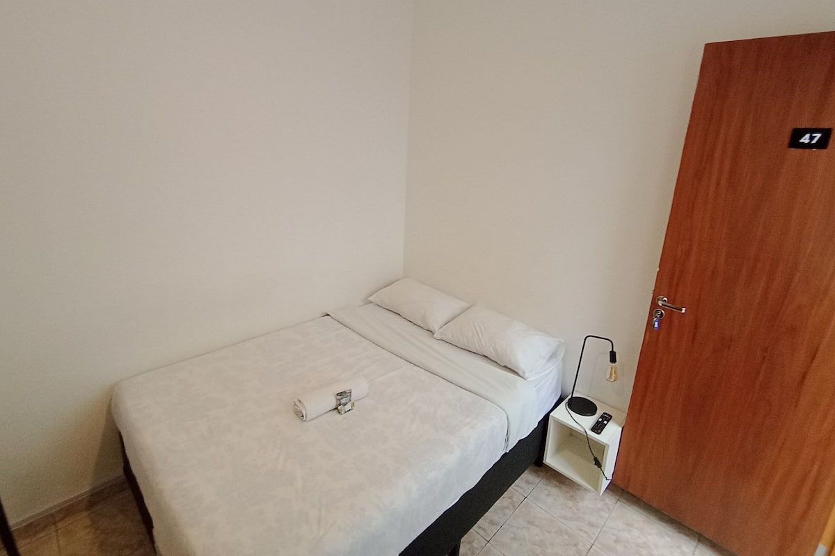 A small, single-bed room with a wooden door marked '47', a bedside table with a lamp, and a power outlet.