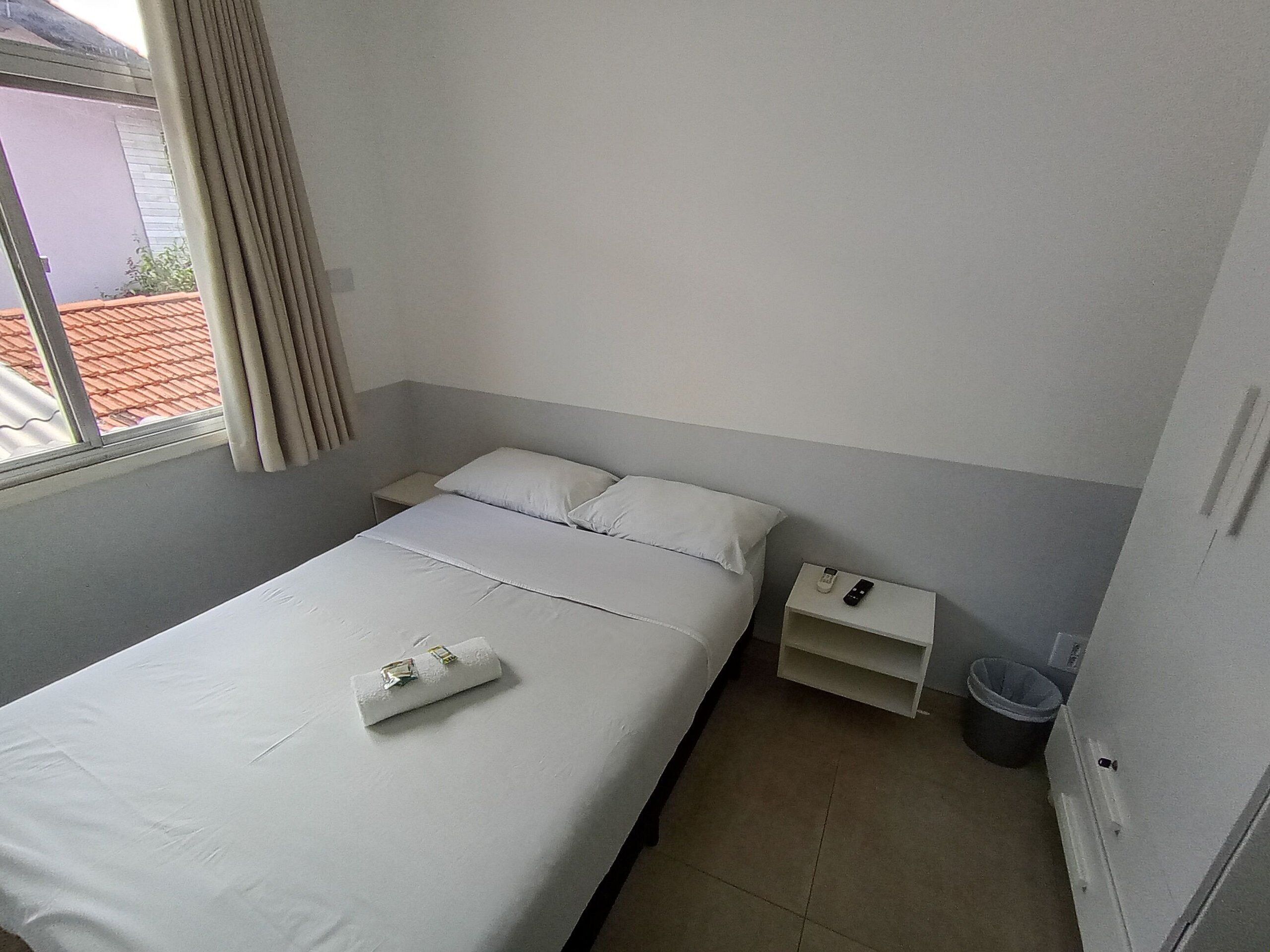 Una cama individual con una almohada, una mesita de noche con un control remoto, un cesto de basura y una ventana con cortinas.