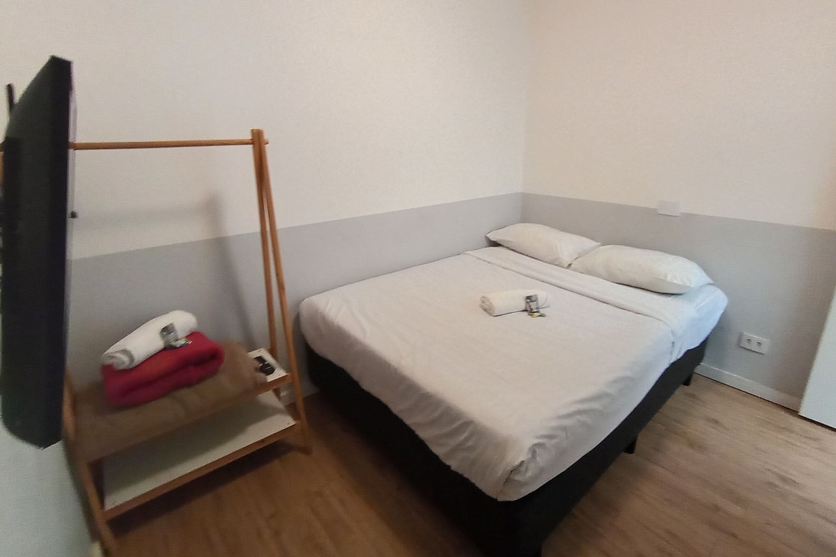 A single bed with white bedding, a wooden shelf with a red towel, and a wall-mounted TV.