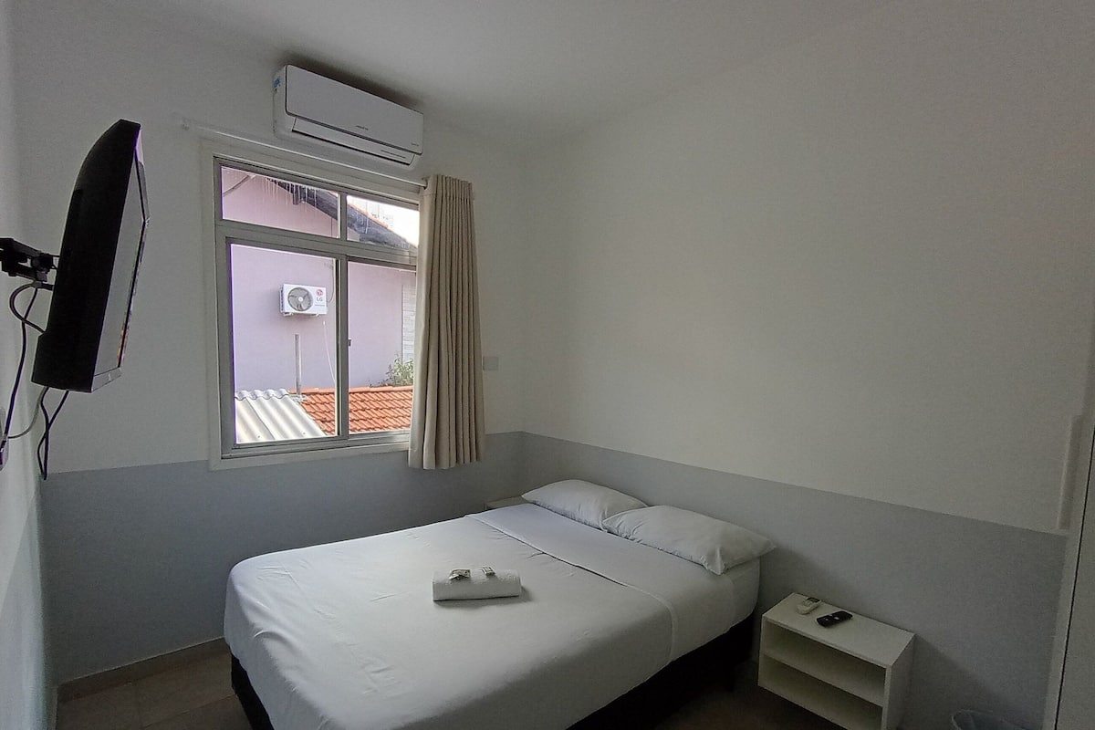 A single-bed room with a window, an air conditioning unit, a TV mounted on the wall, and a bedside table with a remote control.